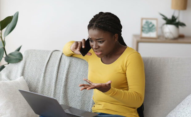 Young black woman on sofa with laptop and cellphone looking frustrated