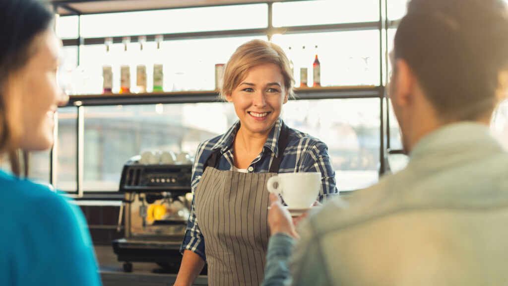 Barista smiling while serving white coffee cup to customer's hand.
